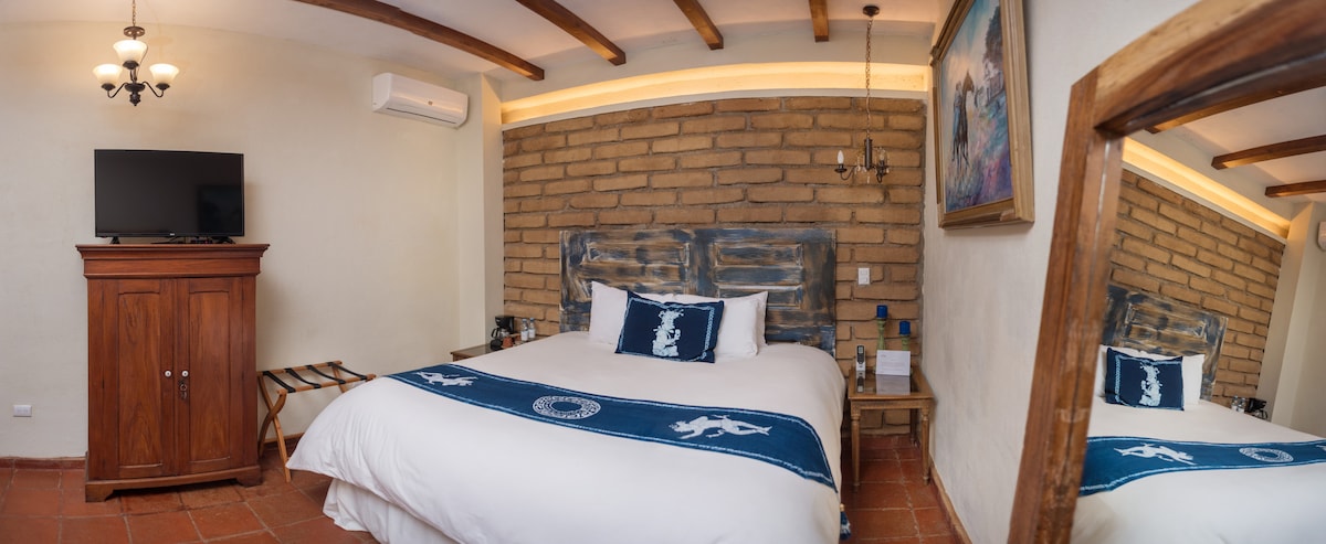 A spacious guest room features a comfortable bed adorned with a blue and white blanket. A flat-screen TV is mounted on the wall, and wooden beams accentuate the ceiling. Natural light illuminates the room, contrasting with the textured brick wall and wooden furniture.