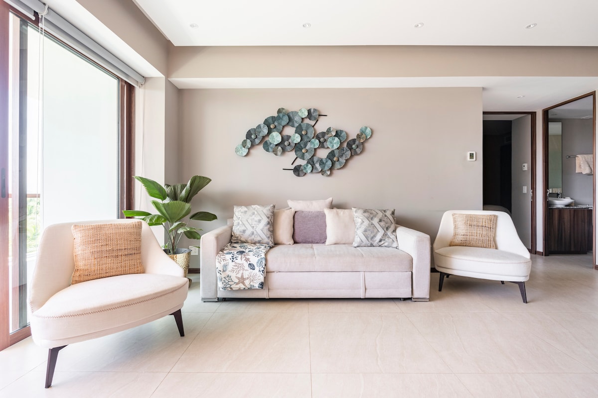 A modern living area features a light-colored sofa adorned with decorative cushions and an elegant piece of wall art. Two stylish armchairs complement the space, while a potted plant adds a touch of greenery. Large windows provide natural light and views of the outside.