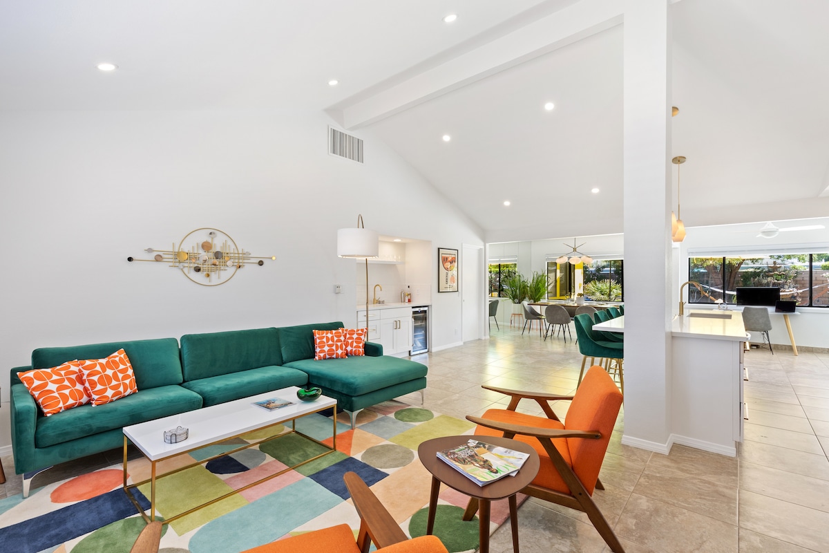 A mid-century modern living area features a large sectional sofa adorned with colorful cushions. A round coffee table sits atop a multi-colored rug. Bright natural light enters through expansive windows, enhancing the airy feel of the open space that connects to the dining area and kitchen.