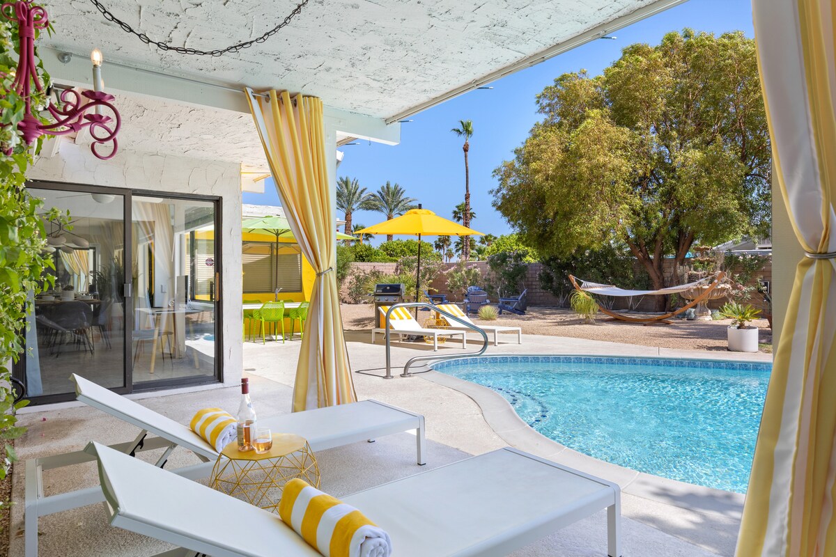A sunlit patio area features white lounge chairs positioned alongside a sparkling pool. Bright yellow umbrellas provide shade, while a hammock is hung in the background. Lush greenery and a lemon tree enhance the serene backyard atmosphere.