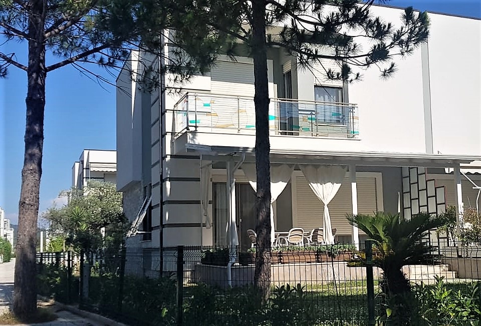 A modern villa is displayed with a two-story design surrounded by greenery. A patio furnished with seating is visible on the upper level, while the ground features a landscaped garden. Large windows allow natural light to illuminate the airy spaces.