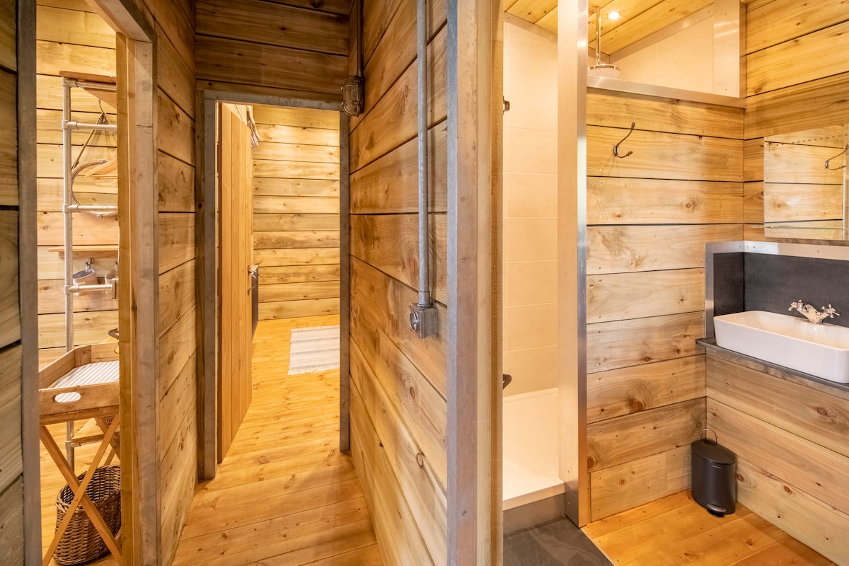 An interior hallway showcases warm wooden walls and flooring. A bathroom area features a modern sink and a shower space. A towel rail and mirror complement the design, while a wooden folding table is visible in the entrance. Soft lighting enhances the natural materials.