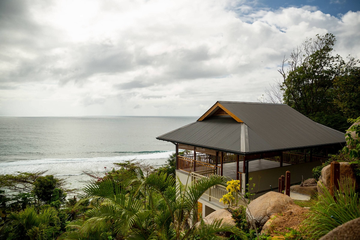 A chalet is situated on a hillside, offering views of the ocean and coastline. Lush greenery surrounds the exterior, while the sloped roof and open terrace provide a welcoming appeal. The sea is visible in the background, with gentle waves lapping against the shore.