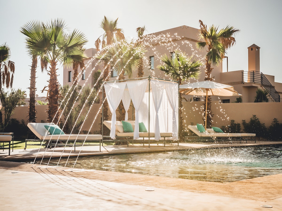 A serene pool area is outlined by lush palm trees and a sunshade. A stylish cabana is draped with white fabric, while sun loungers with teal cushions invite relaxation. Water sprays gracefully from elegant fountains, glistening in the sunlight.