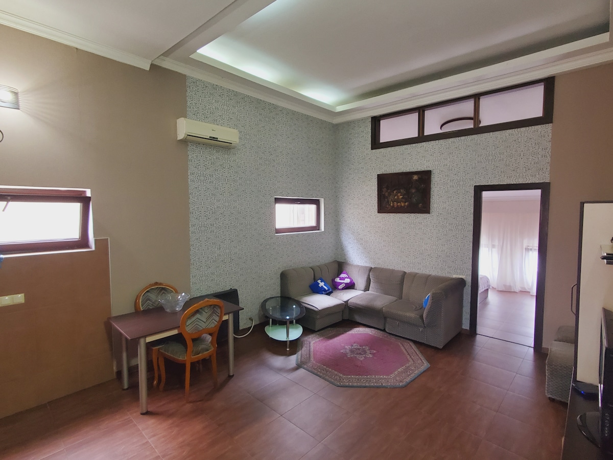 A living area features a gray sectional sofa and a small glass coffee table. A simple dining table with two wooden chairs is positioned against the wall. Natural light enters through a window, highlighting patterned wallpaper and a decorative wall piece.