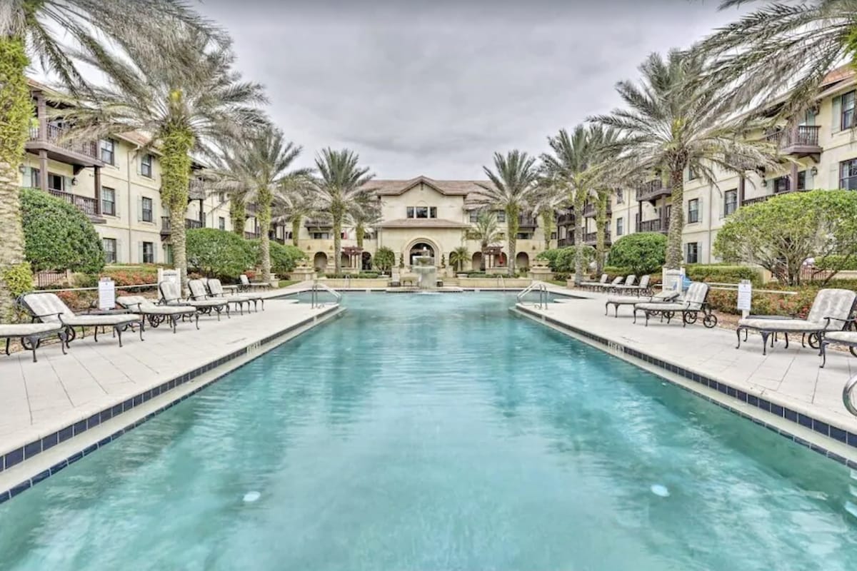 A tranquil resort-style pool is framed by palm trees and lounge chairs on either side. The water features a clear blue hue, reflecting the surrounding architecture and greenery. The pool area is complemented by an elegant structure in the background, enhancing the peaceful atmosphere.