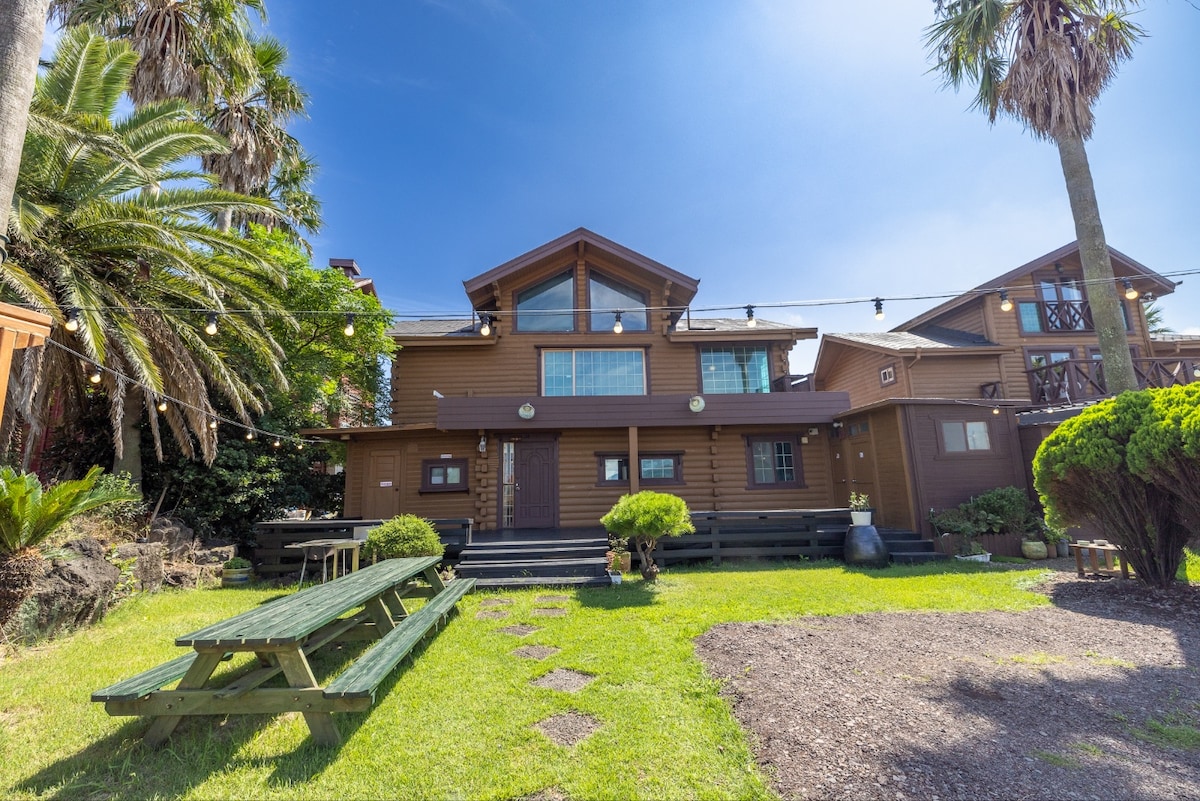 A spacious outdoor area is featured, surrounded by lush greenery and palm trees. Multiple picnic tables are arranged on a well-maintained lawn, leading to a two-story wooden structure with ample windows and exterior lights, indicative of a welcoming atmosphere.