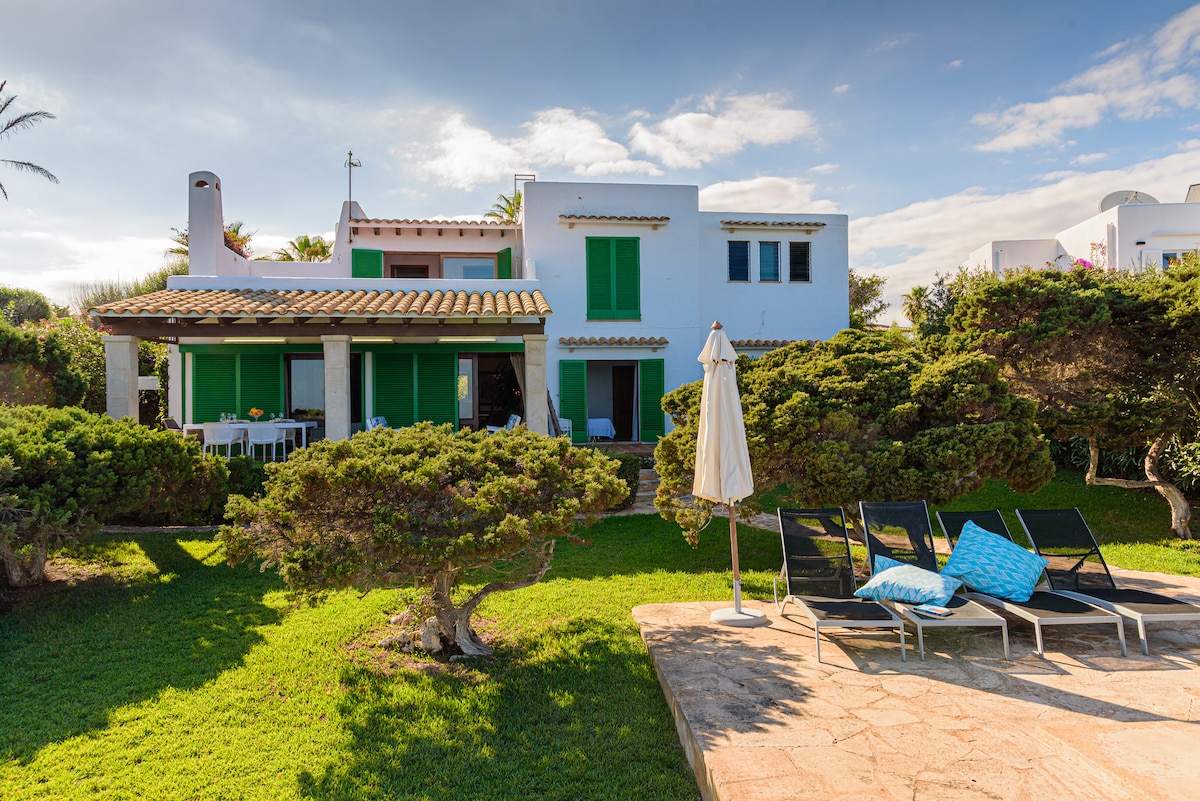 The exterior of the villa features a lush green lawn and well-maintained shrubs. The building showcases white walls with green shuttered windows. A patio area with lounge chairs and a sun umbrella is visible, providing a relaxing space to enjoy the outdoors.