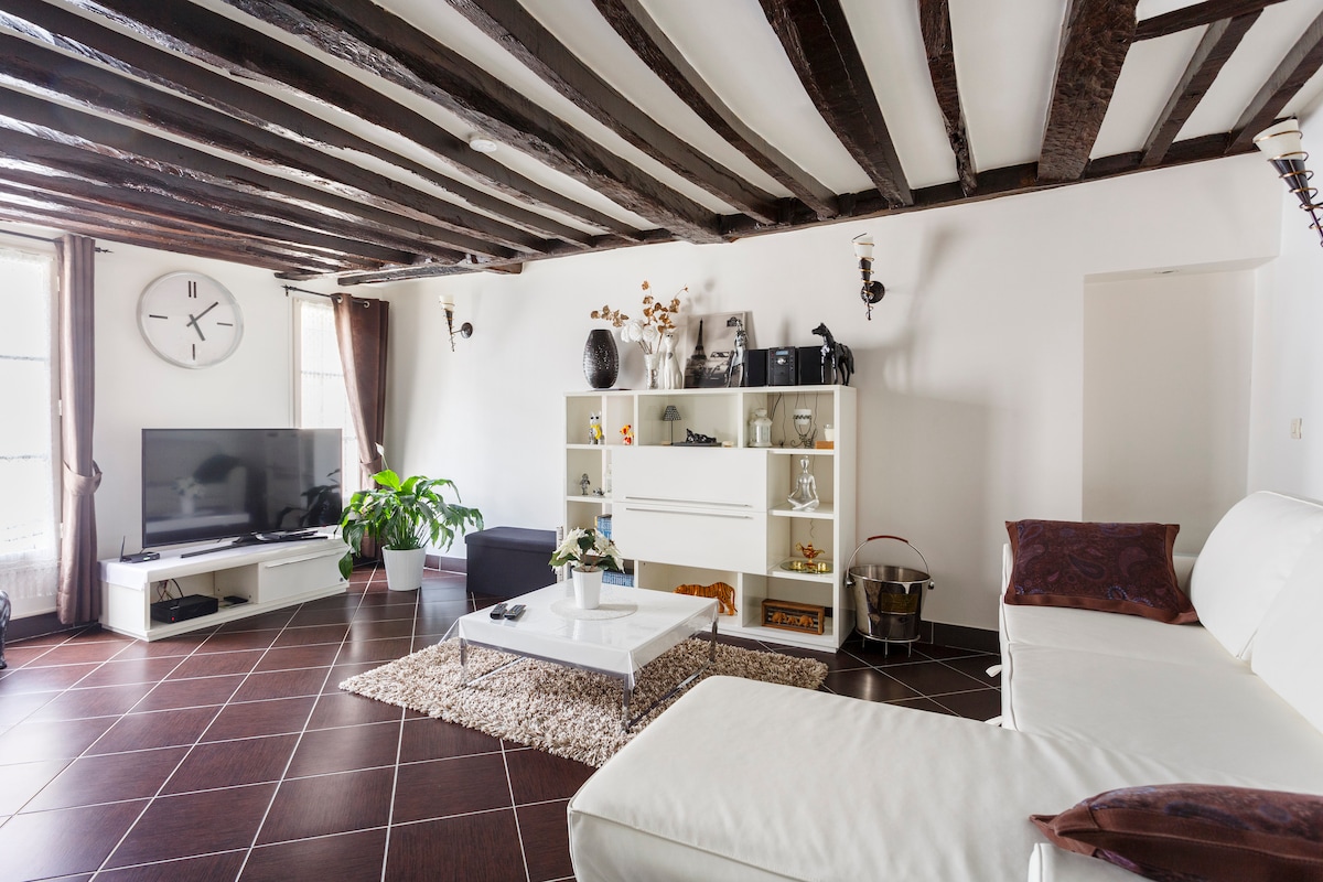 A spacious living area is highlighted by exposed wooden beams and a large flat-screen television. A white sectional sofa and coffee table are positioned on a textured rug. Decorative items and a potted plant add a touch of warmth to the modern design.