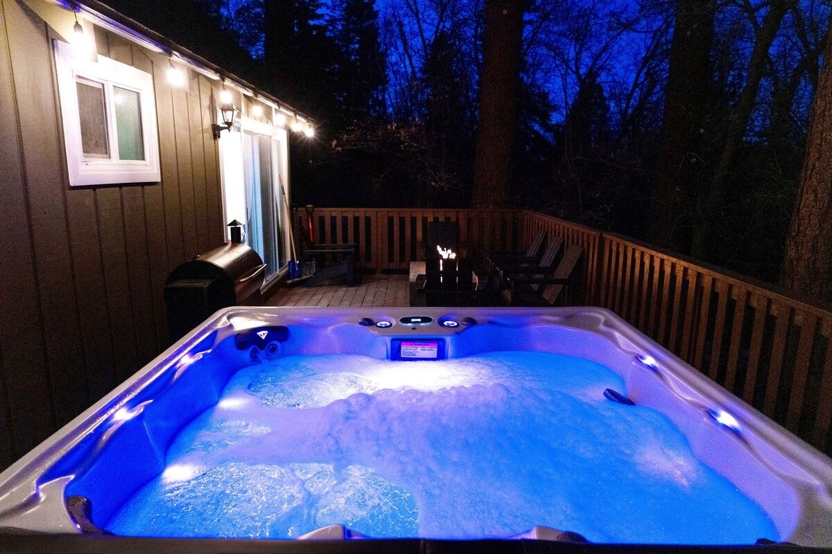 The outdoor patio features a large jacuzzi illuminated with soft blue lights, surrounded by wooden decking. Cozy seating is provided nearby, along with a barbeque grill. The tranquil setting is set among towering trees, enhancing the serene atmosphere in the evening.