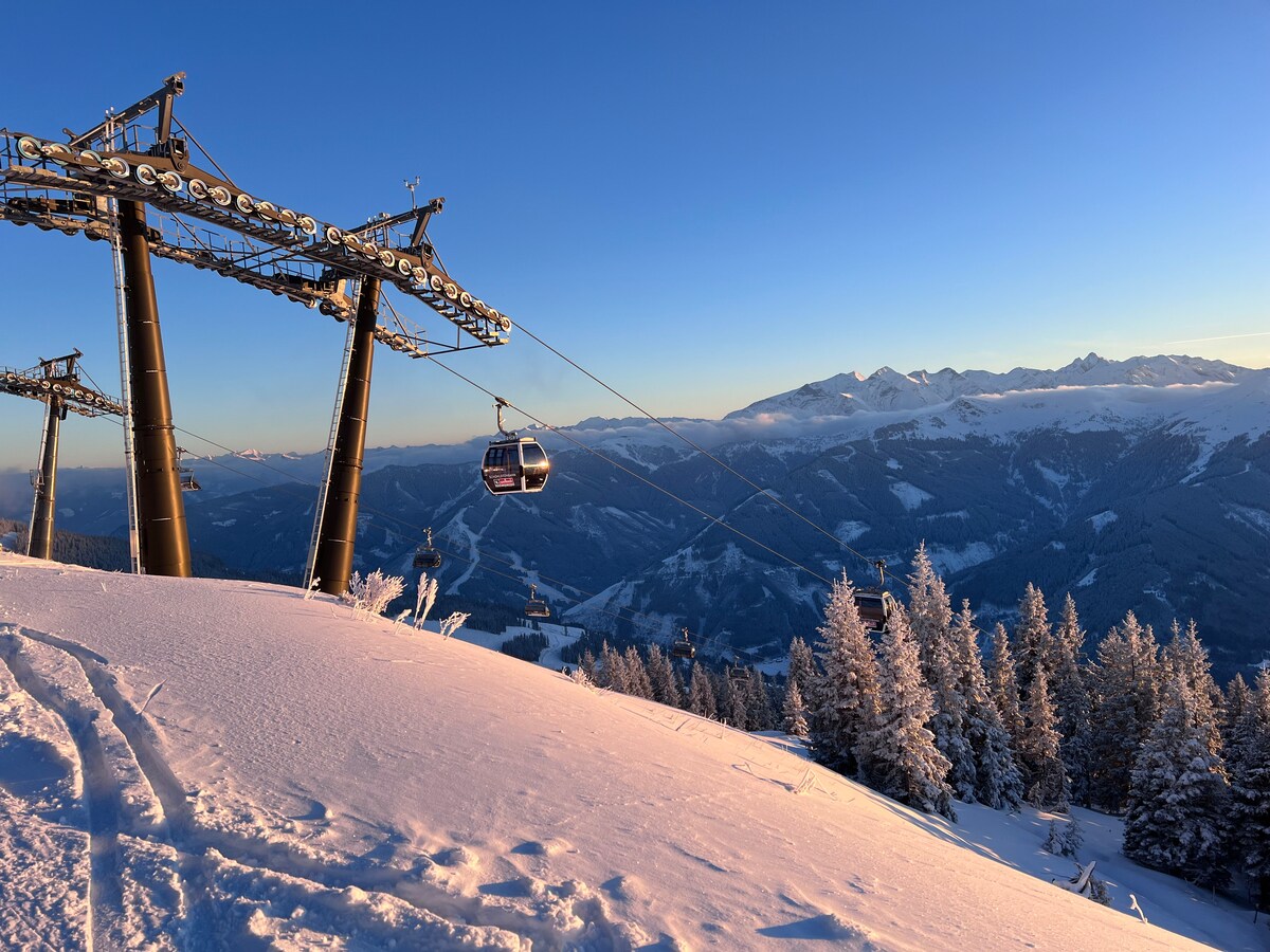 A ski lift ascends over a snowy landscape, with a gondola in transit against a backdrop of distant mountains and a clear blue sky. Sunlight casts a warm glow on the snow-covered ground, highlighting the evergreen trees blanketed in white.