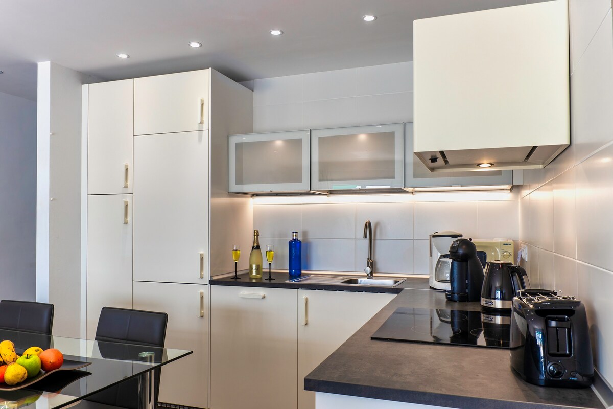 A modern kitchen is equipped with an array of appliances, including a coffee maker, kettle, and wine bottles on the countertop. White cabinets and sleek surfaces are complemented by under-cabinet lighting, enhancing the clean lines of the space. A glass dining table with a fruit bowl is nearby.