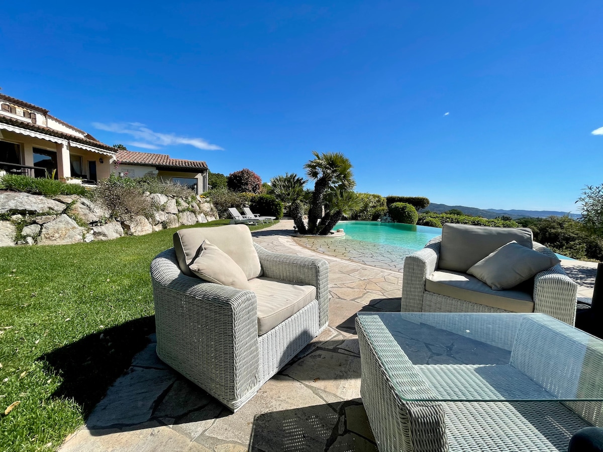A furnished outdoor seating area features two cushioned chairs and a glass-top table, set against a backdrop of a landscaped garden and a pool. The sky is clear and blue, while palm trees provide natural texture to the serene space.