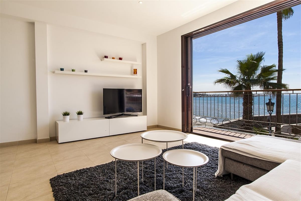 A modern living space features a large flat-screen TV positioned on a white media unit. Three round coffee tables are set on a textured rug. Double glass doors open to a balcony with views of the sea and a palm tree outside.