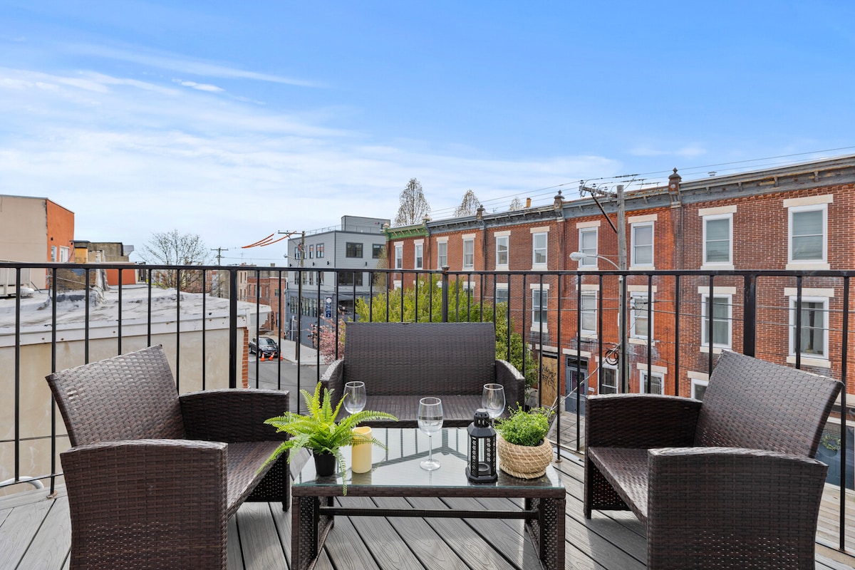 The private rooftop deck features a glass-top table surrounded by four woven chairs, offering a space for relaxation. A small potted plant and decorative lantern are placed on the table, while the urban skyline and colorful buildings are visible in the background.