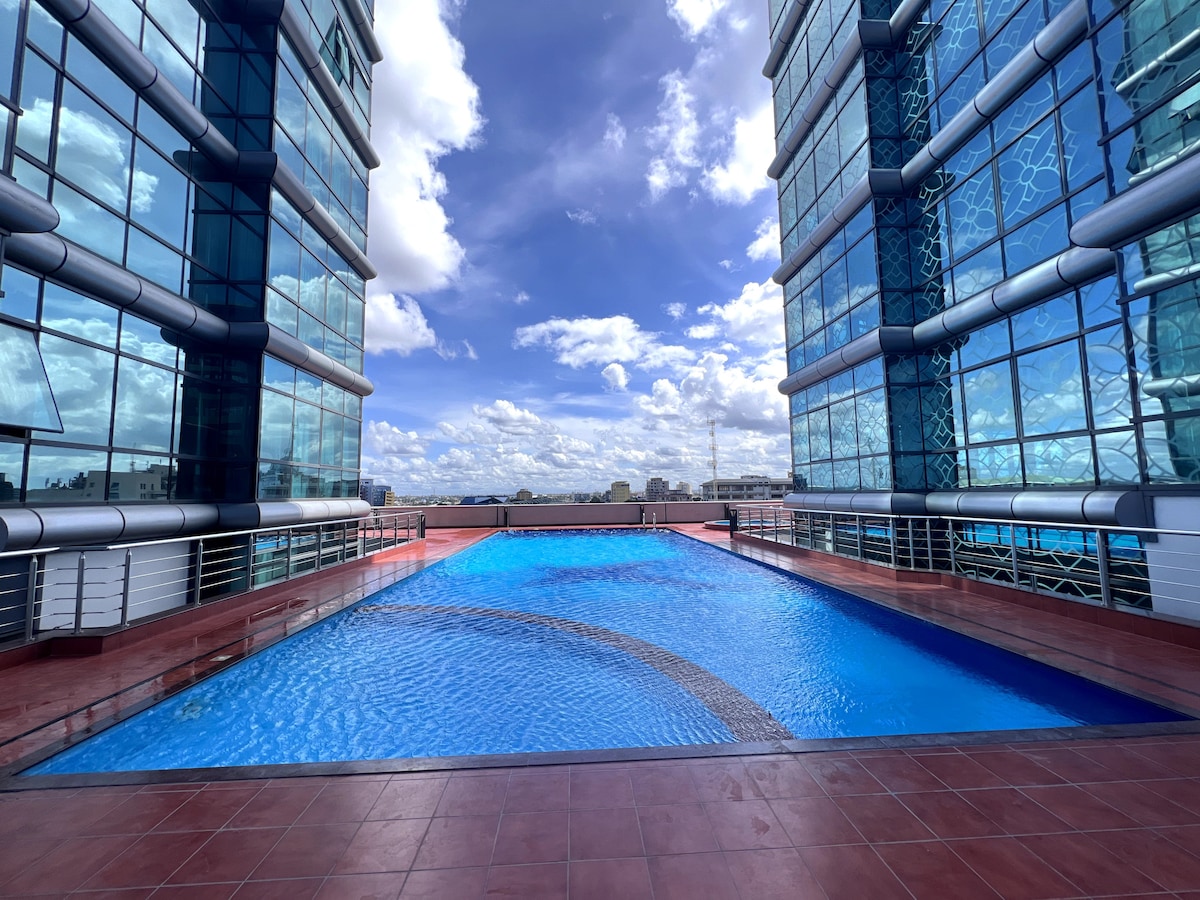 An Olympic-sized swimming pool is set between two modern glass buildings, reflecting the clear blue sky. The pool area features a spacious deck with sleek, red-tiled flooring, providing a serene environment for relaxation and recreation.