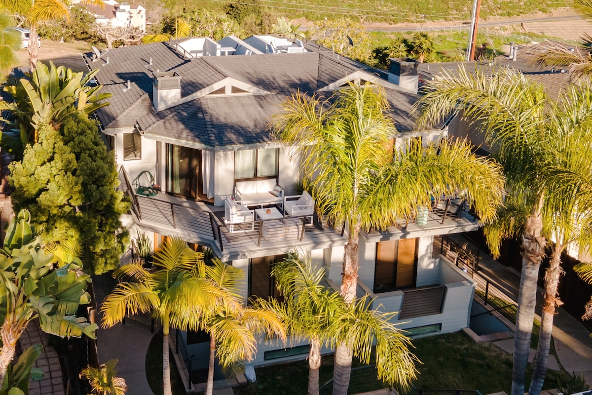 An aerial view captures the contemporary home surrounded by lush palm trees. A spacious deck is visible, furnished with seating, while the roof showcases a modern design. The neighborhood features greenery and open spaces, contributing to a serene setting.