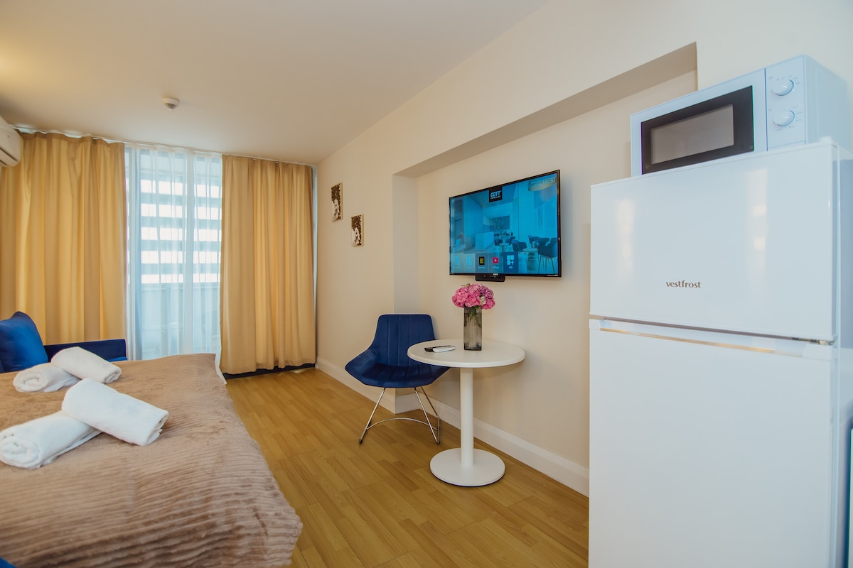 A cozy living space features a spacious bed with neatly arranged towels. A small white table with blue chairs is positioned beside a refrigerator and microwave. Light streams in through large windows covered by soft curtains, creating a bright and inviting atmosphere.