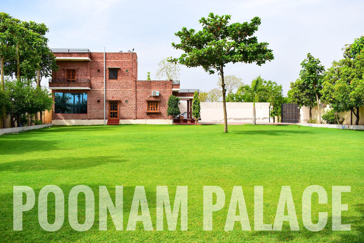 A traditional villa, Poonam Palace, is set against a backdrop of lush green grass. The structure features a brick façade and large windows, allowing natural light to fill the interiors. Surrounding trees provide shade, enhancing the serene outdoor environment.