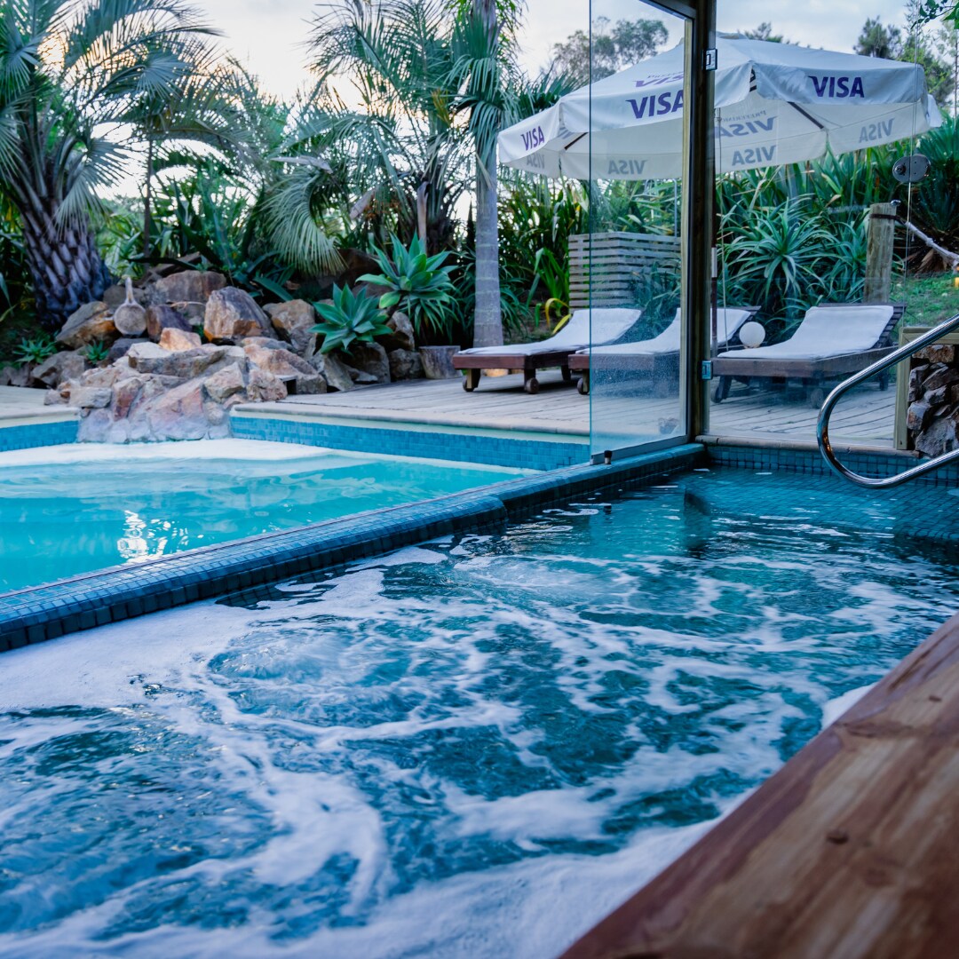 A tranquil spa area features a hydrojet pool and a separate swimming pool, surrounded by lush tropical plants. Comfortable loungers are positioned nearby, and a shaded area offers protective umbrellas. The serene atmosphere invites relaxation and connection with nature.