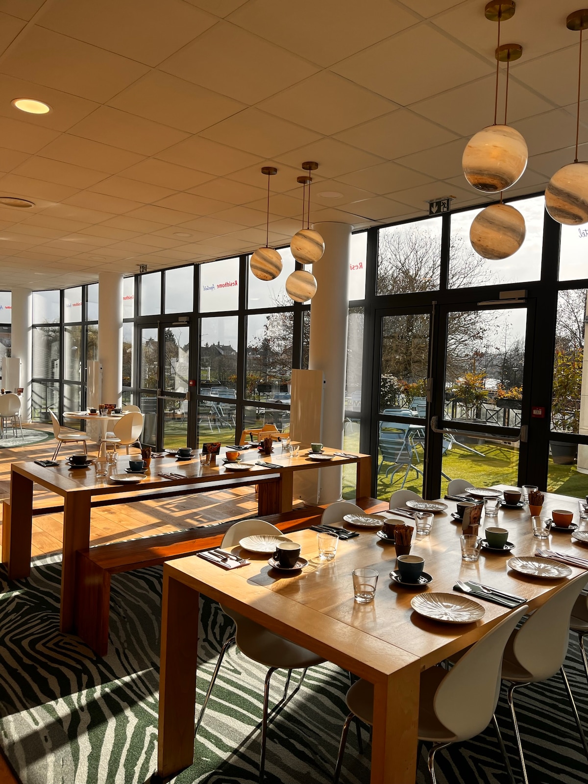 A spacious dining area is presented, featuring wooden tables arranged for dining. Place settings with plates and cups are visible at each table. Large glass doors allow natural light to flood the room, with a view of greenery seen outside.