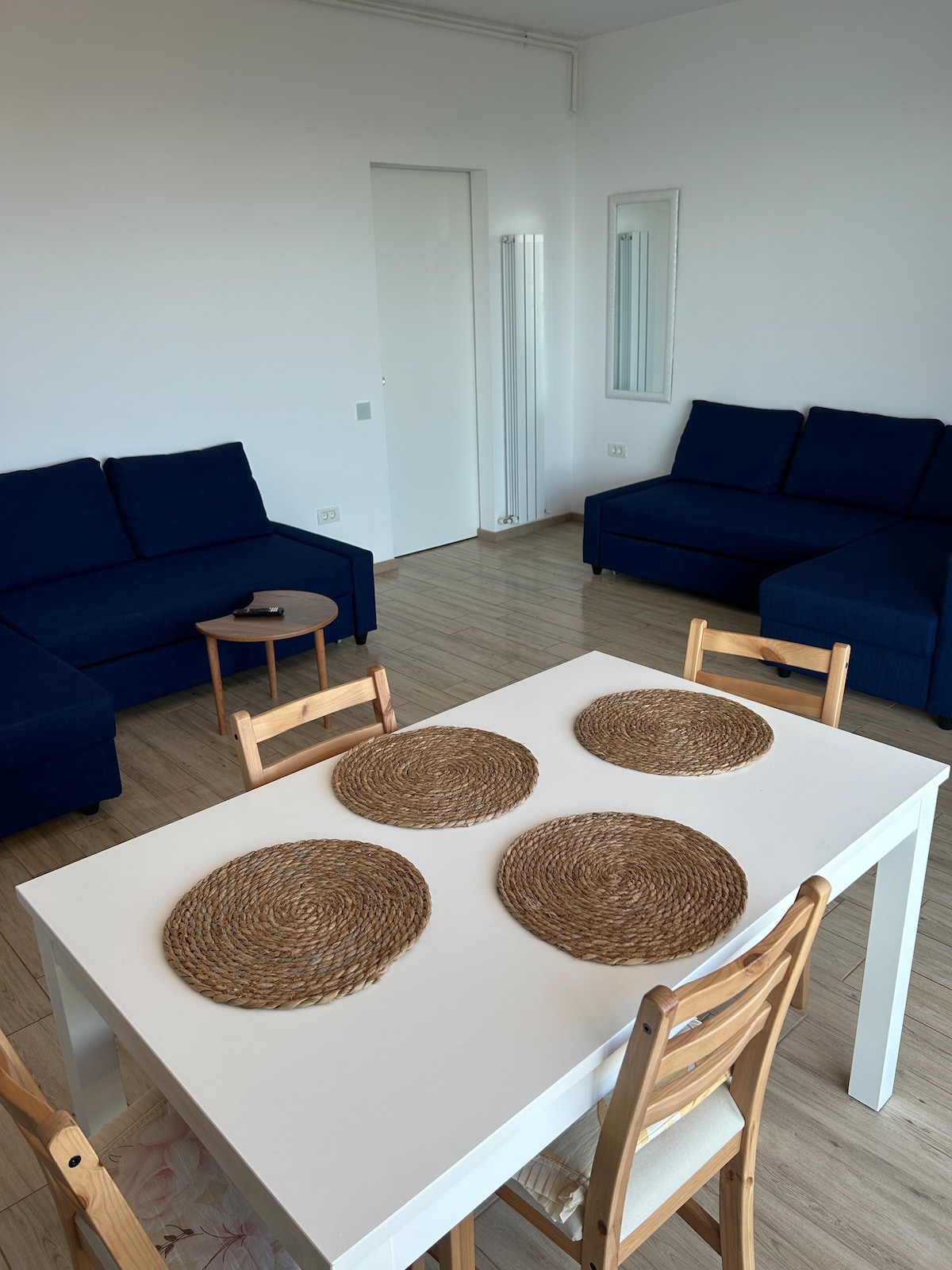 A spacious living area features two dark blue sofas positioned near a small round table. A white dining table set with four woven placemats is visible, providing a simple and functional dining space. Light natural wood flooring complements the bright atmosphere.