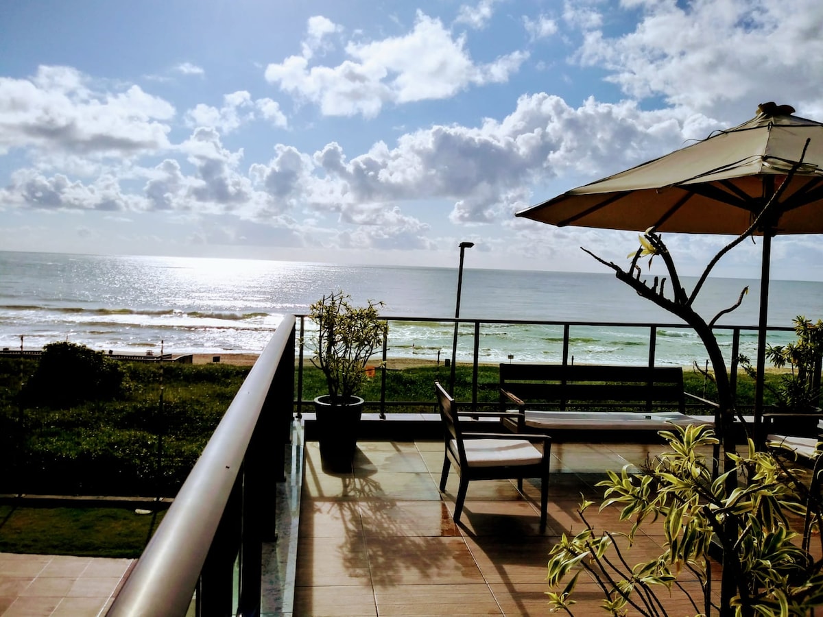 A spacious terrace offers a scenic view of the ocean, with gentle waves lapping at the shore. Sunlight reflects off the water, while a few plants and a seating area with a sun umbrella are arranged on the balcony, inviting relaxation.