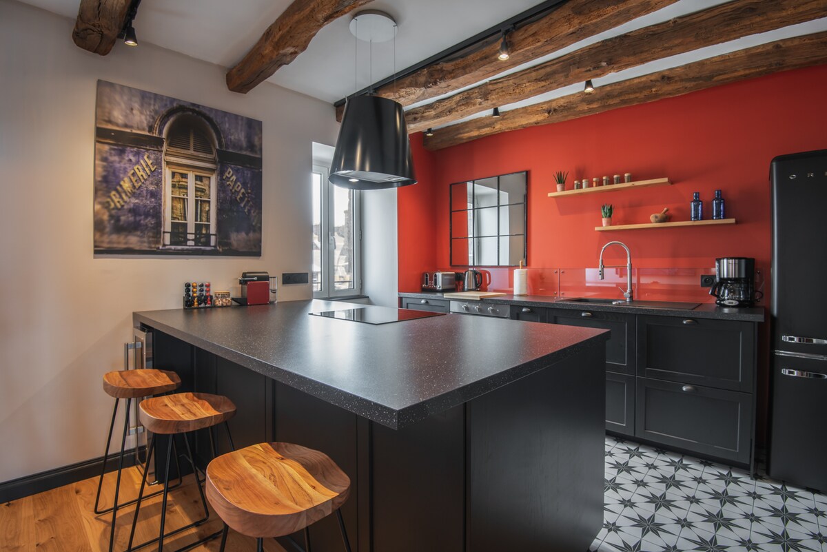A modern kitchen features a large black island with four wooden stools. Red and black cabinets provide storage, while a window allows natural light to fill the space. A coffee machine and kettle are visible on the countertop, framed by decorative plants on the shelves.