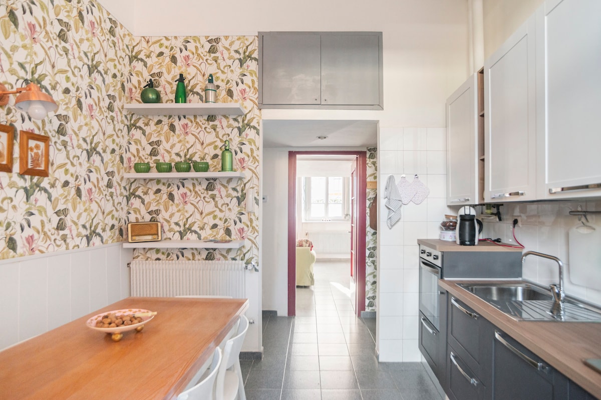 A modern kitchen is furnished with a wooden dining table and white chairs. Open shelves display green dishware against floral wallpaper. A sink and stove are positioned along the dark cabinetry. The space transitions to a living area visible through an adjacent doorway.