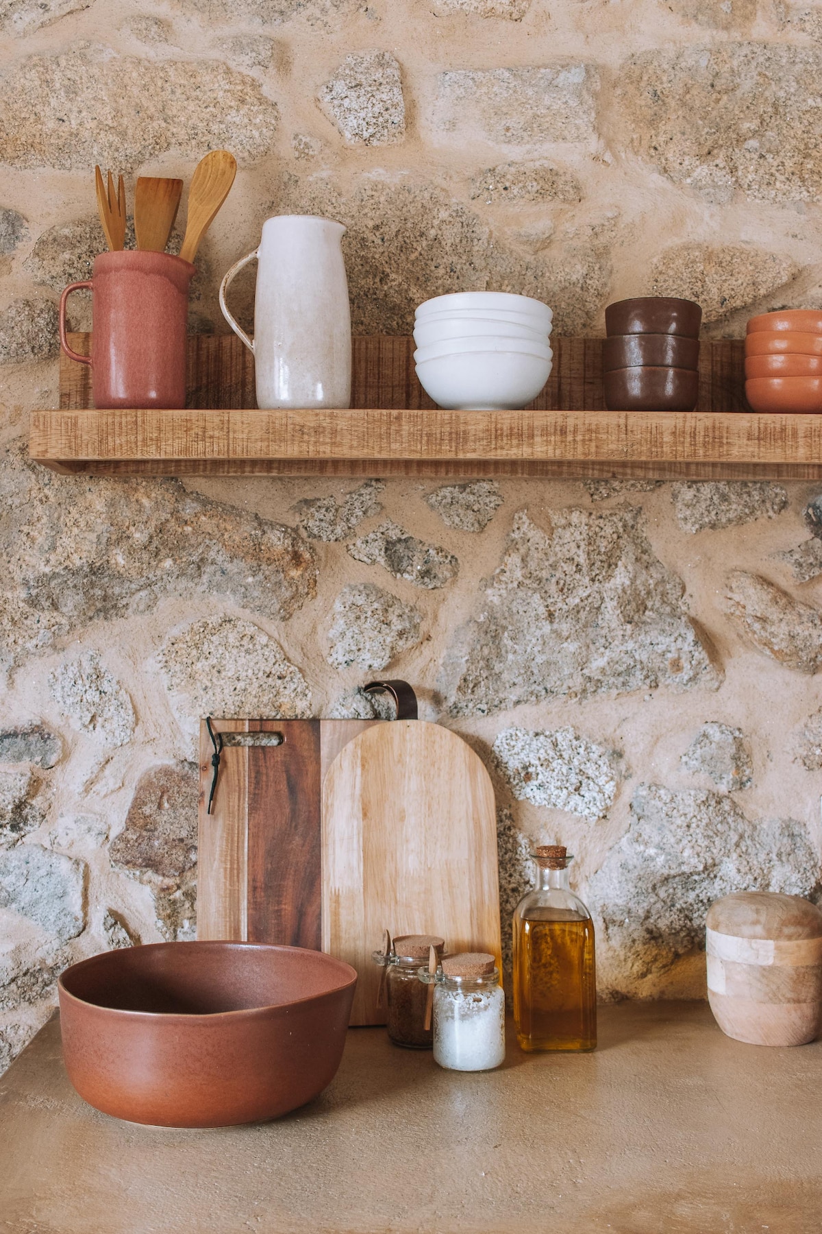 A wooden shelf holds various ceramic cups and utensils, complemented by glass and clay containers. Below, a smooth stone countertop features a rustic bowl, a wooden cutting board, and a glass bottle filled with oil, creating a warm and functional kitchen setting.