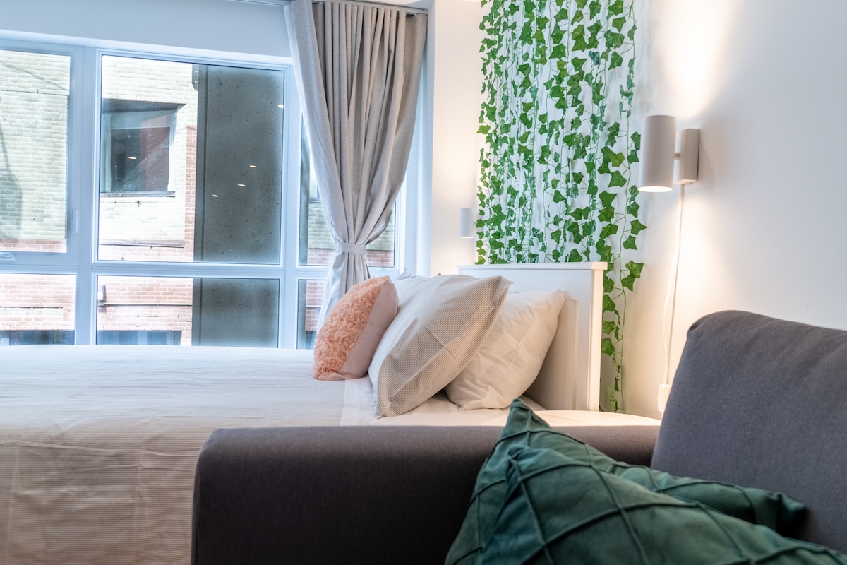 A cozy bedroom is displayed, featuring a spacious bed draped in light-colored linens. A gray sofa adds comfort to the space. Green ivy decorates the wall, while large windows allow ample natural light, framed by soft curtains.