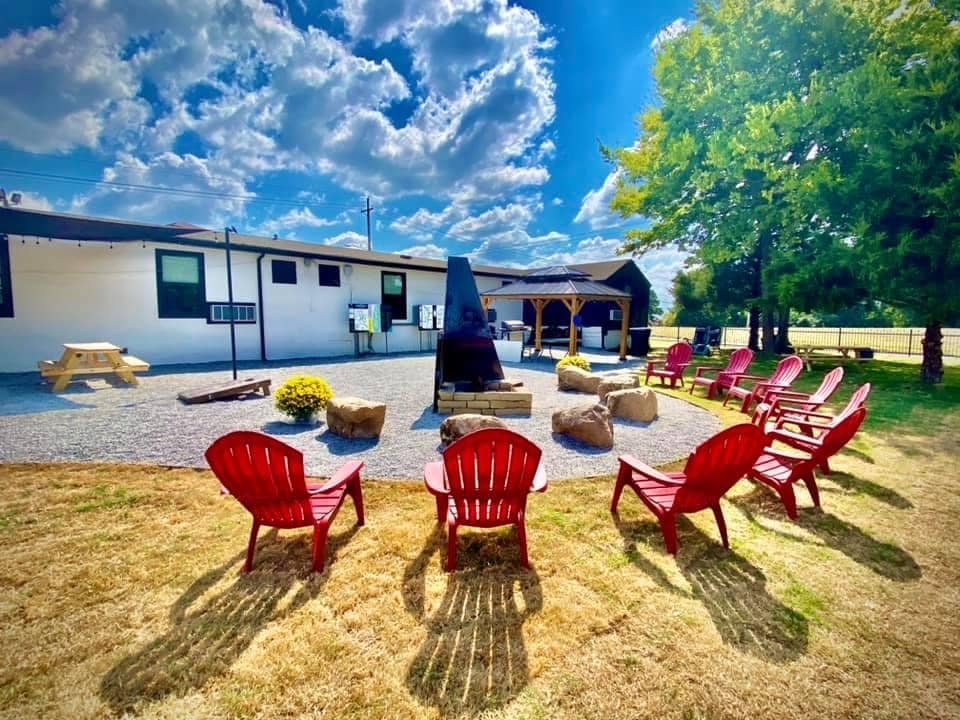An outdoor gathering space is showcased, featuring a circular arrangement of red Adirondack chairs surrounding a central fire pit. Nearby, a wooden picnic table and a gazebo are visible, framed by a grassy area and dotted with manicured flower beds under a bright sky.