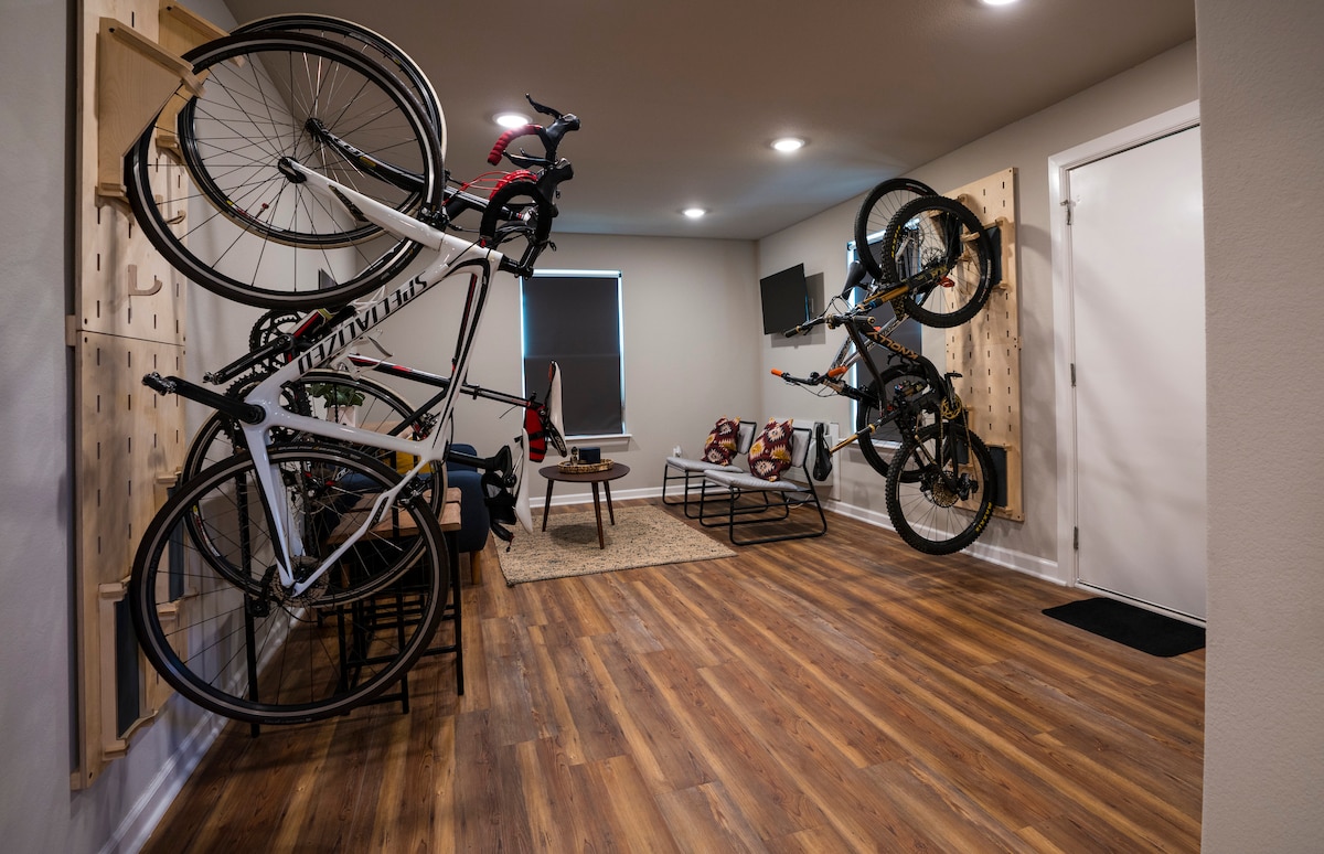 A spacious area is visible with wooden floors and bright overhead lighting. Two bicycles are securely mounted on the wall. A low coffee table sits on a textured rug, surrounded by colorful seating. A window allows natural light to fill the room.