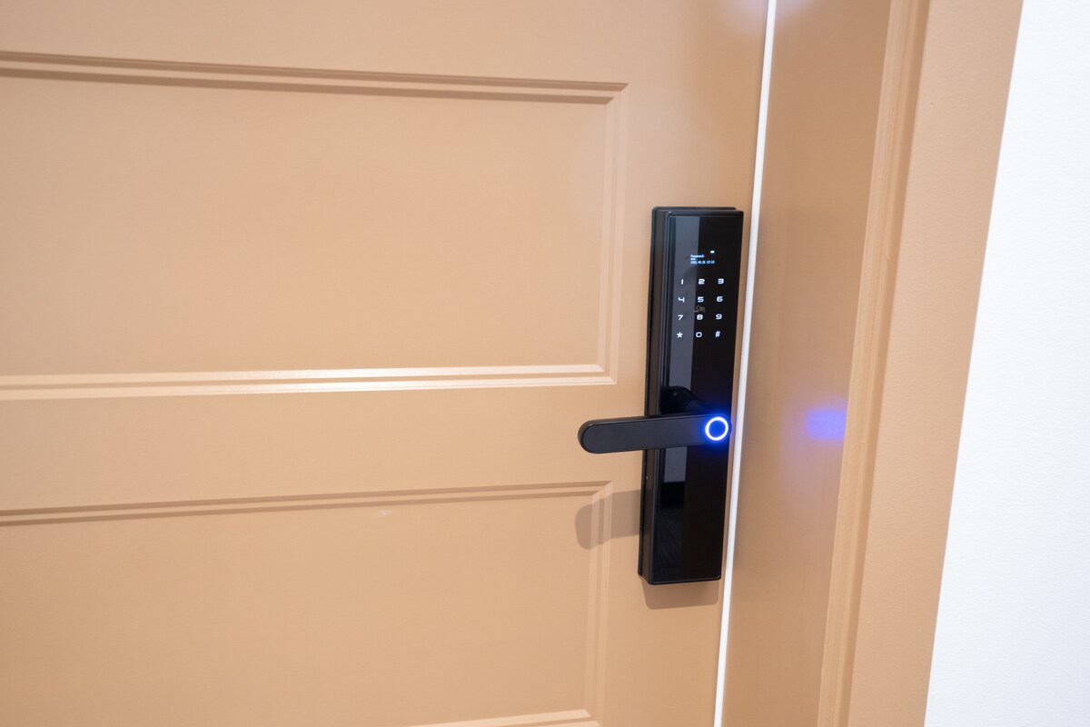 A modern keyless entry system is installed on a sleek, tan door. The lock features a touchpad with backlit numbers and a circular LED indicator that emits a soft glow. The door's smooth surface and clean lines contribute to the suite's contemporary aesthetic.