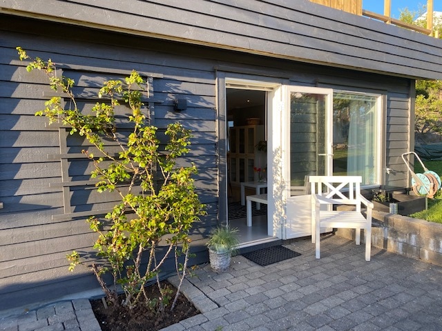 The exterior entrance to the property is highlighted by a small patio area featuring a white chair and a potted plant. The facade consists of dark wooden panels, and a large glass door provides access to the interior.