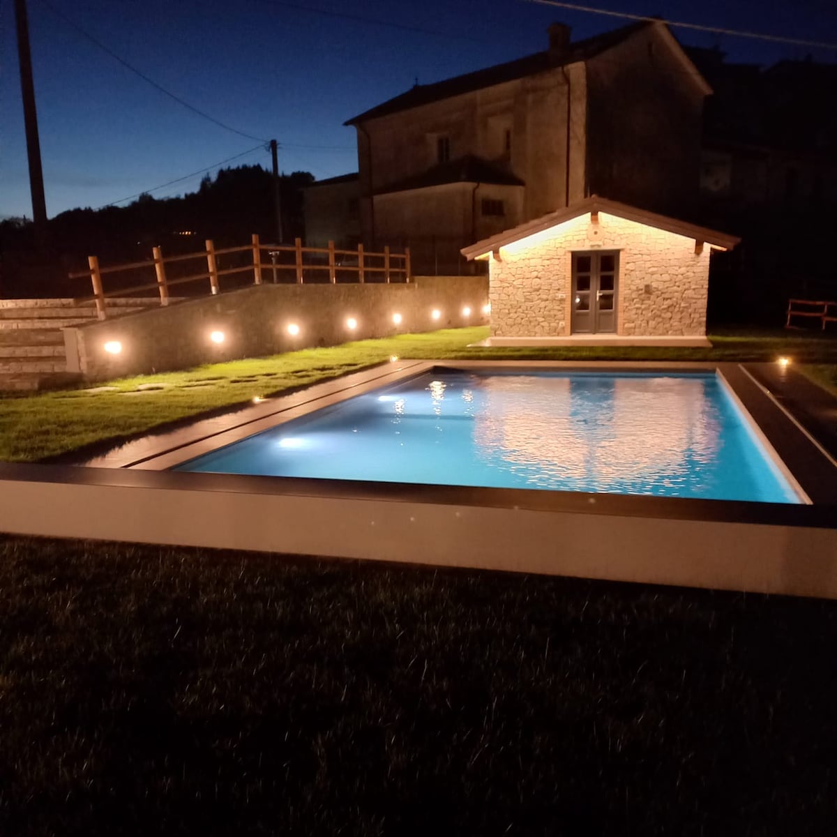 A well-lit swimming pool is set within a grassy area, reflecting the evening sky. A small wooden structure is visible nearby, featuring large glass doors. The surrounding space is illuminated by soft lighting, creating a serene ambiance.