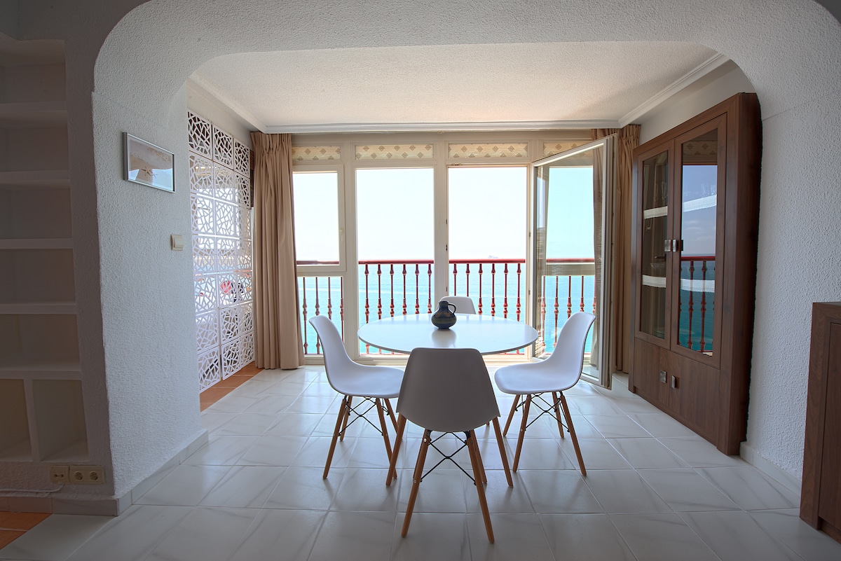 A dining area with a round table surrounded by four white chairs is displayed. The large windows open to reveal views of the sea, allowing natural light to fill the space. Decorative details on the walls add an elegant touch.