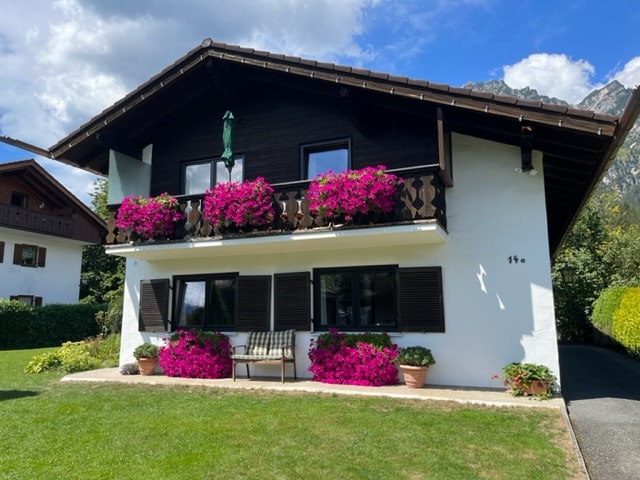 A charming two-story building features a dark wood exterior with wooden shutters. Bright pink flowers are positioned in window boxes and planters against a backdrop of lush green grass. The surrounding landscape includes trees and mountains visible in the distance under a partly cloudy sky.