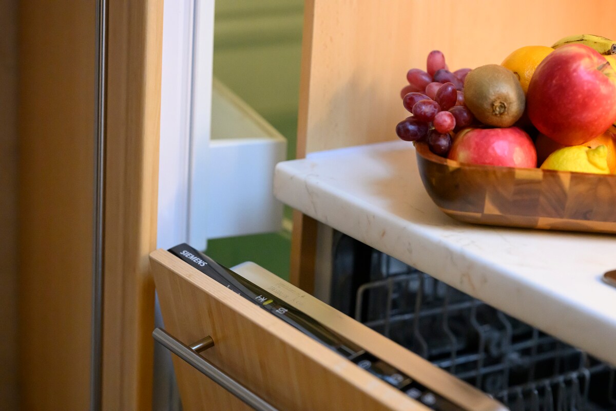 A wooden bowl filled with fresh fruits, including grapes, apples, and citrus, sits atop a marble surface. The image also features the edge of a modern dishwasher, adding to the practical kitchen design.
