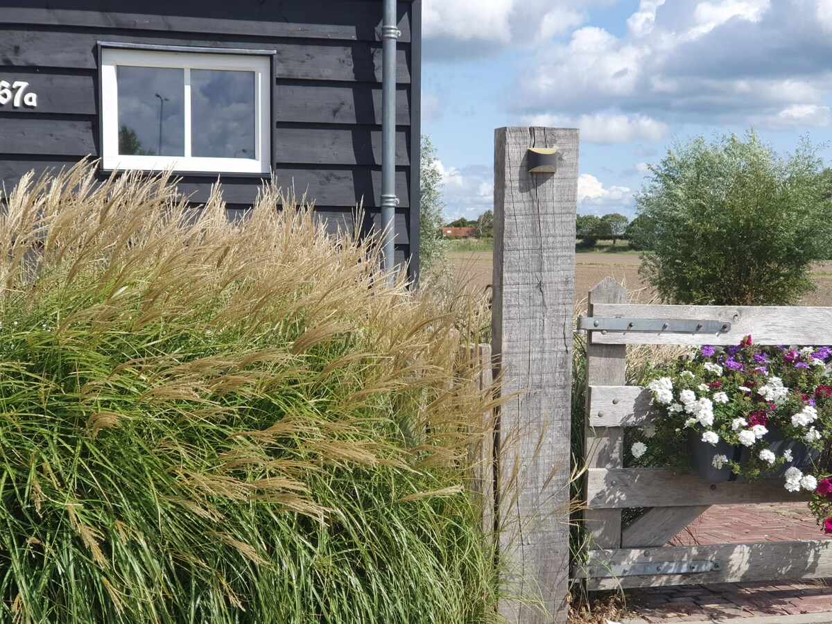 Tall grass sways gently in front of a dark wooden building, featuring a partially visible window. A rustic wooden fence, adorned with bright flowers in hanging boxes, creates a welcoming entrance to the property. Open fields stretch in the background under a partly cloudy sky.