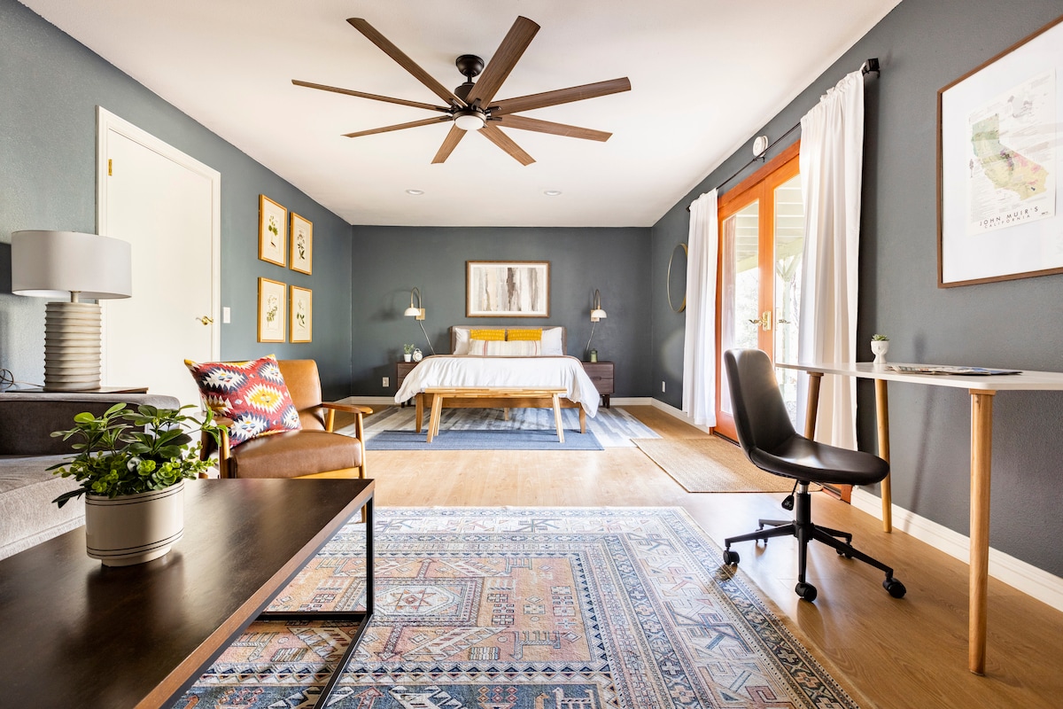 A spacious guest suite is highlighted by a queen bed with a light duvet and decorative pillows. Natural light enters through double doors, leading to an outdoor area. A ceiling fan and warm wood accents create a comfortable atmosphere, complemented by a small desk and seating area.