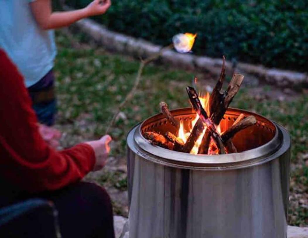 A fire pit is centered in a grassy area, surrounded by stones. Several logs are arranged to create a lively flame. One person, seated and holding a stick, prepares to roast a marshmallow, while another stands in the background, adding to a cozy outdoor atmosphere.