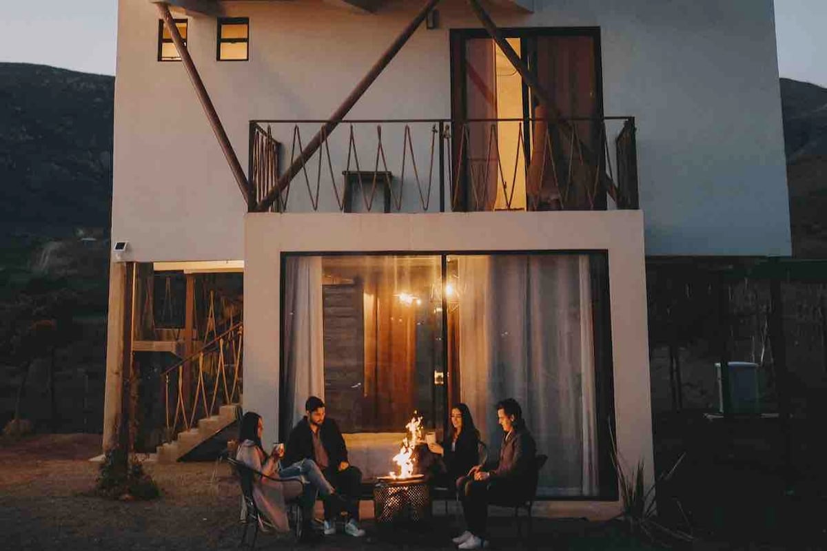 An exterior view of a modern retreat at dusk, showcasing a fire pit surrounded by five seated individuals. The building features a balcony with a railing and large windows that allow soft light to illuminate the space. The surrounding landscape includes distant hills.