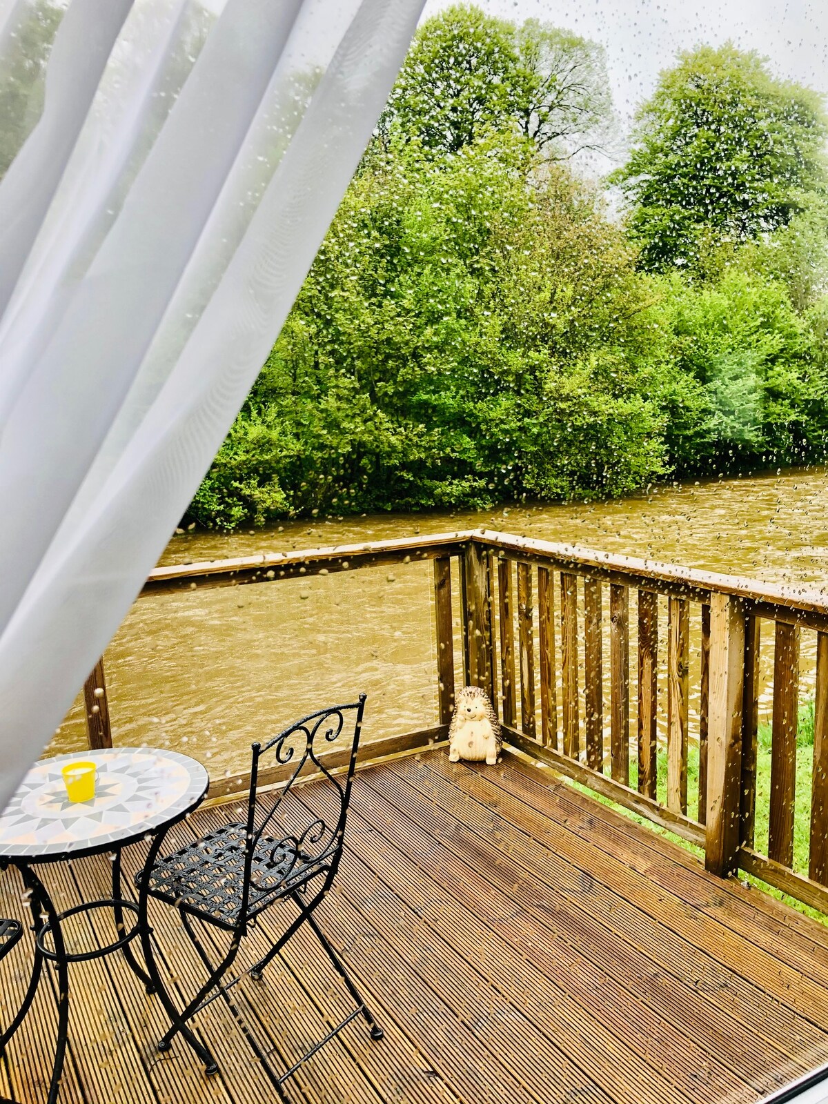 A view from the veranda shows a table and two chairs positioned on the wooden deck, overlooking the flowing River Teviot. Raindrops are visible on the window, with greenery framing the scene, enhancing the serene atmosphere.