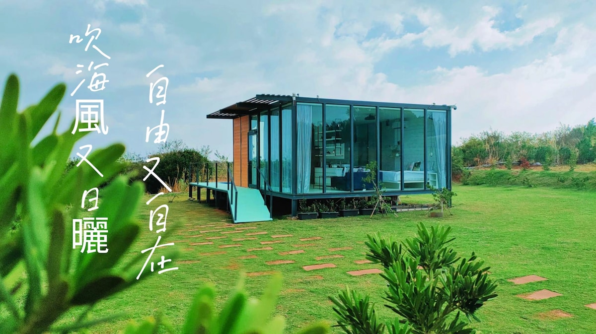 A modern glass structure is set on a lush green expanse. The building features large windows and a vibrant blue interior, allowing natural light to flood the space. Surrounding greenery enhances the serene atmosphere, complemented by a clear sky above.