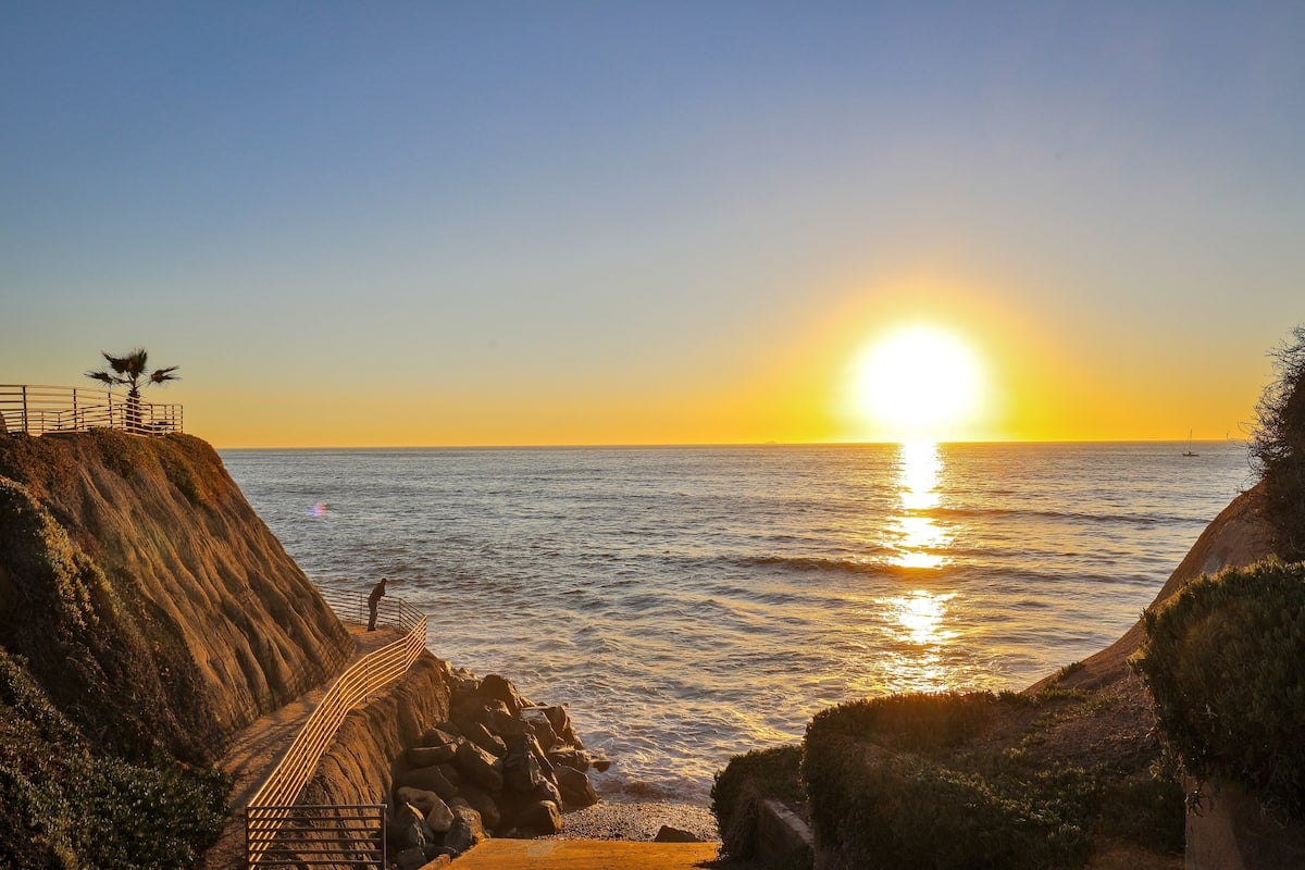 A serene coastal view is presented, showcasing the sun setting over the ocean. Silhouetted cliffs frame the scene, while a wooden walkway leads toward the water. Gentle waves reflect the golden hues of the sunset, creating a peaceful atmosphere.