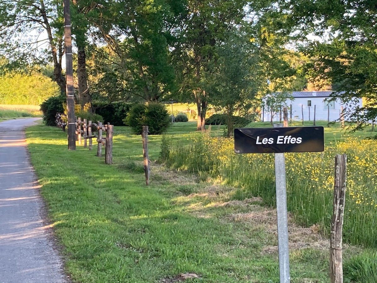 A sign indicating 'Les Effets' is positioned along a grassy pathway, lined with wooden posts. Lush trees and flowering plants enhance the natural surroundings, while a modest building is partially visible in the background, framed by greenery.