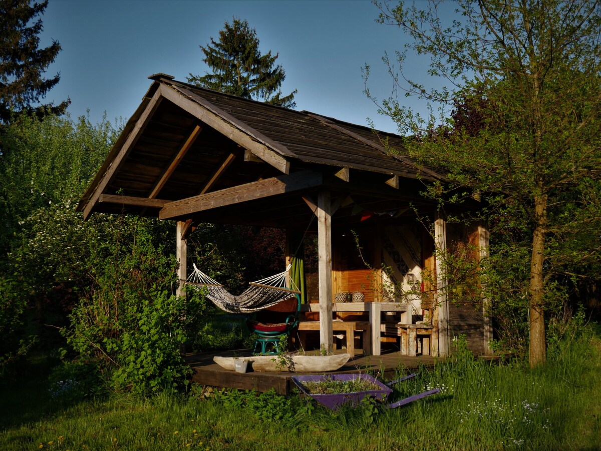 A wooden structure with a sloped roof is surrounded by greenery, featuring a hammock, a table, and chairs underneath. Natural light casts warm tones on the wooden surfaces, while trees and foliage frame the scene, creating a serene outdoor setting.