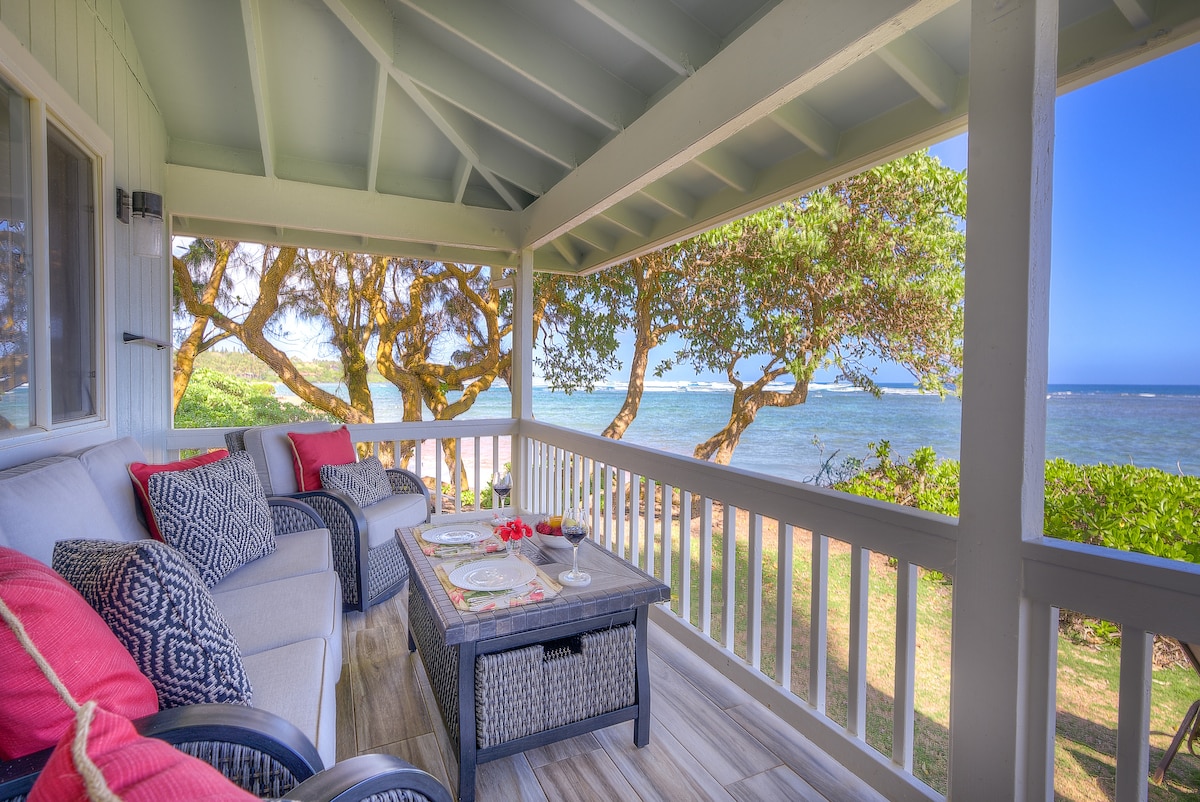 A covered lanai offers seating on a comfortable sofa and armchairs, adorned with colorful cushions. A low table is set with refreshments. Lush trees frame the view of the ocean and shoreline, bringing the natural beauty of the surroundings into focus.