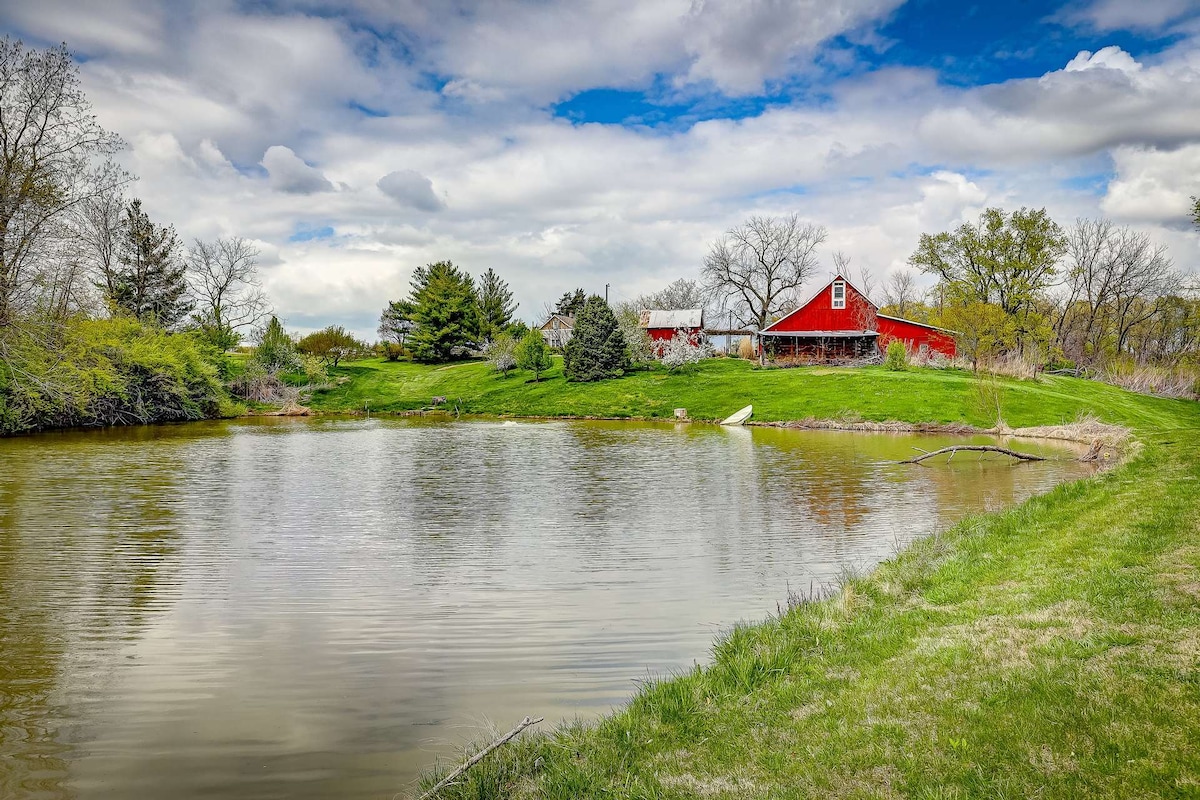 A tranquil pond is situated in the foreground, surrounded by lush greenery and trees. A red barn is visible in the background against a cloudy sky, creating a serene rural landscape. The setting conveys an inviting atmosphere for relaxation and outdoor activities.